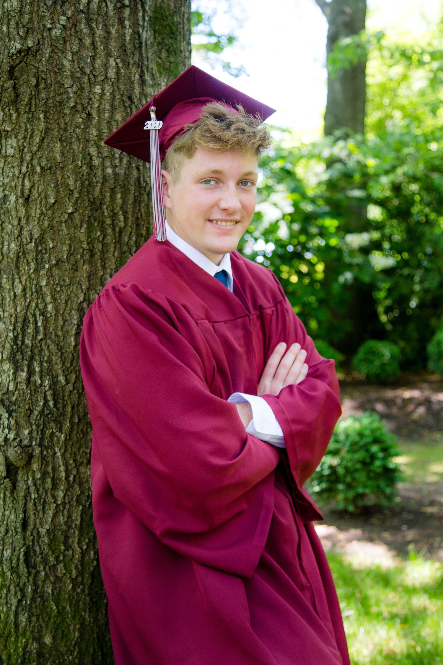 Male graduate in burgundy cap and gown leaning against tree with arms crossed.