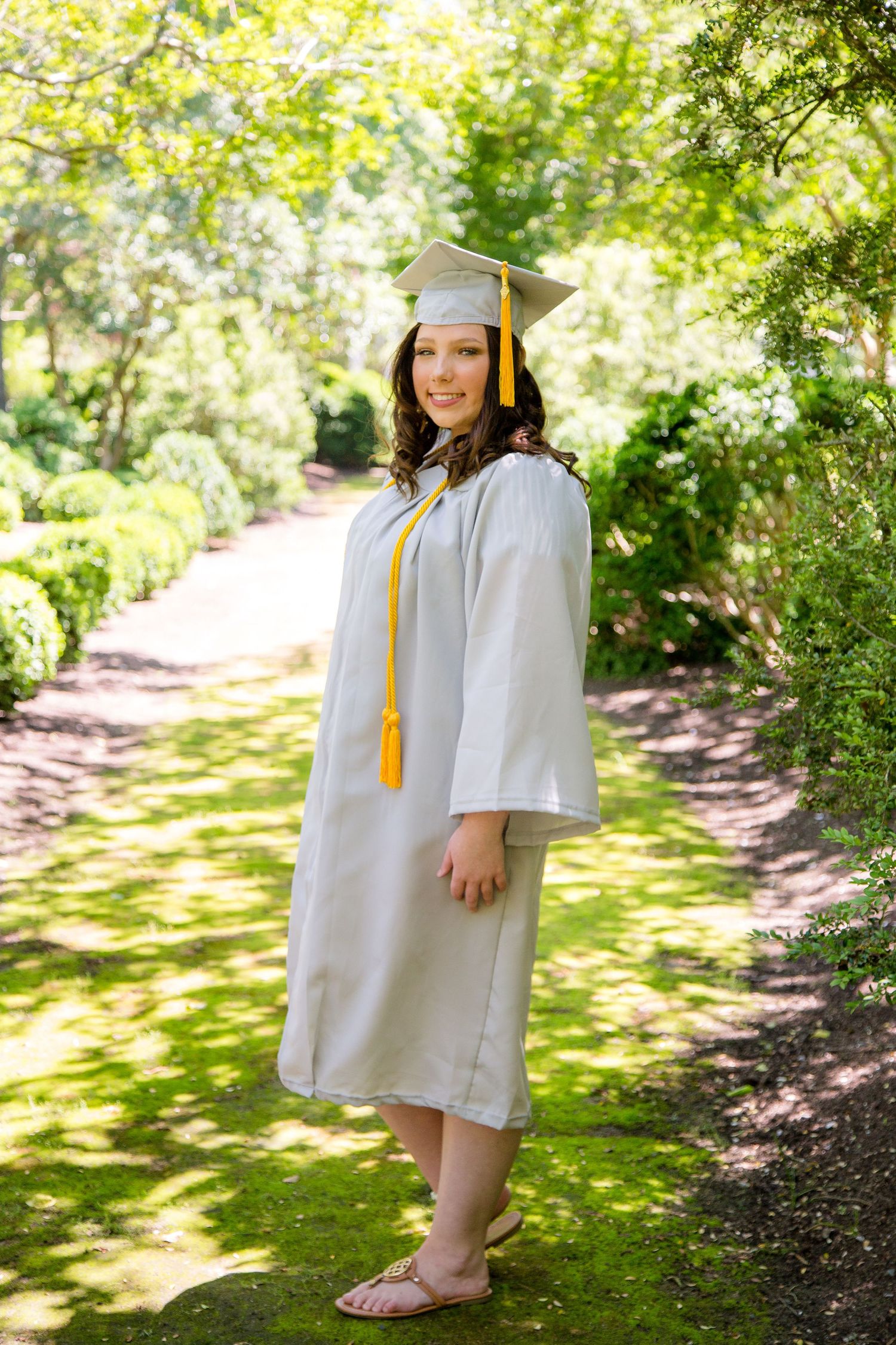 Female graduate in gray cap and gown with yellow tassel standing on garden pathway.