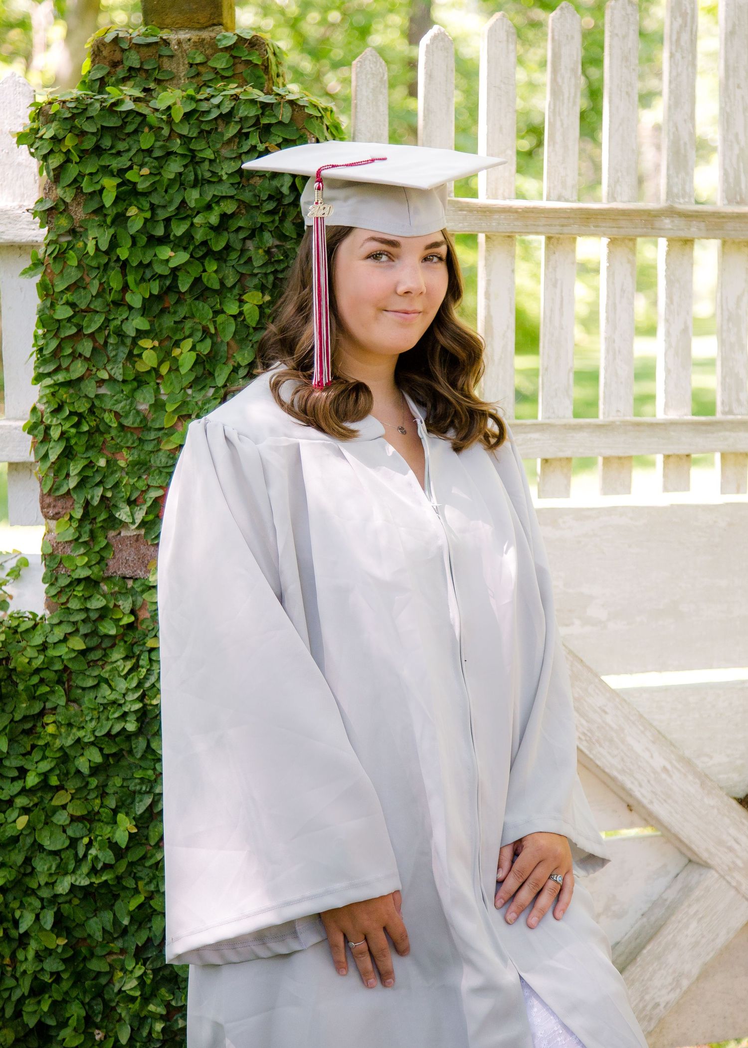 Female graduate in white cap and gown with red tassel posing by ivy-covered fence.