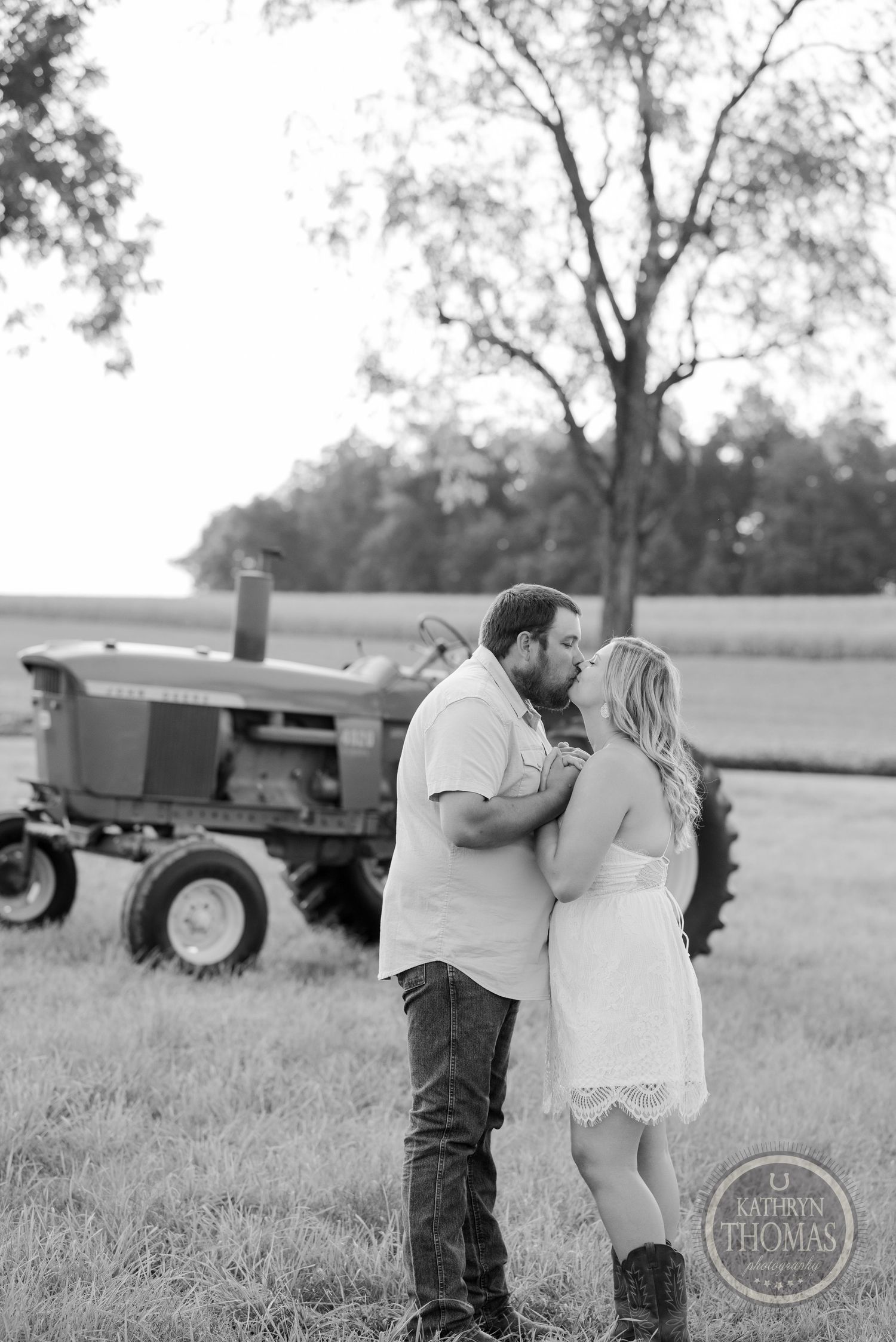 Black and white portrait of couple embracing by vintage tractor in countryside setting.