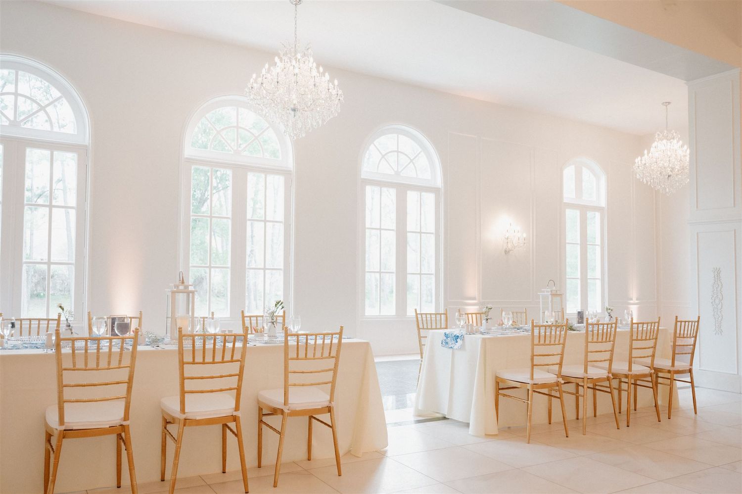 Bright white ballroom with arched windows, crystal chandeliers, and gold chiavari chairs.