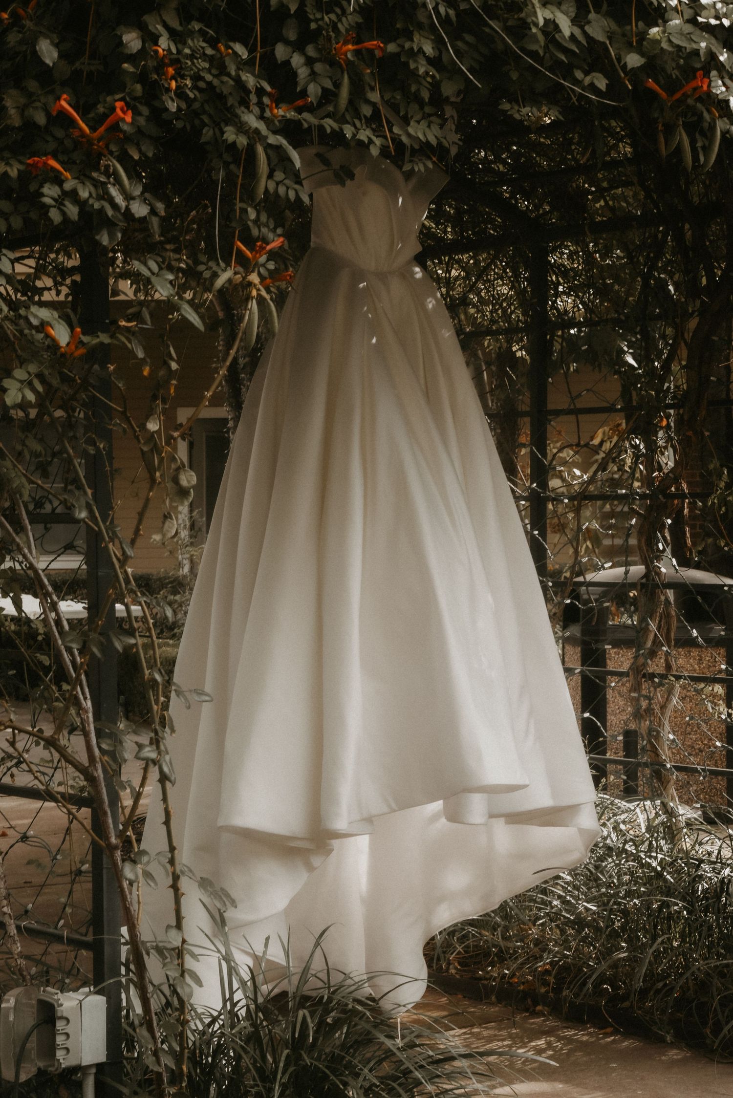 A flowing white wedding dress displayed against dark greenery and brick wall.