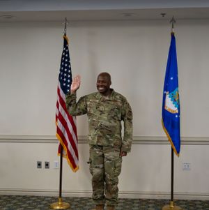 A U.S. service member in military fatigues stands at attention between American and Air Force flags.