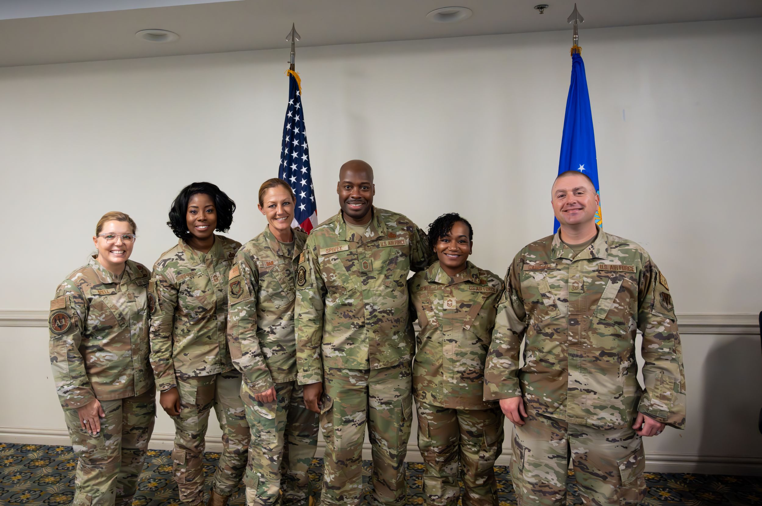 Military personnel pose for a group photo between American and Air Force flags in an indoor setting.