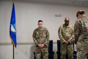 Military service members gather for a ceremony in front of a blue Air Force flag.