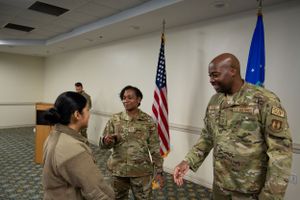 Service members interact during a casual meeting in a conference room with flags displayed.