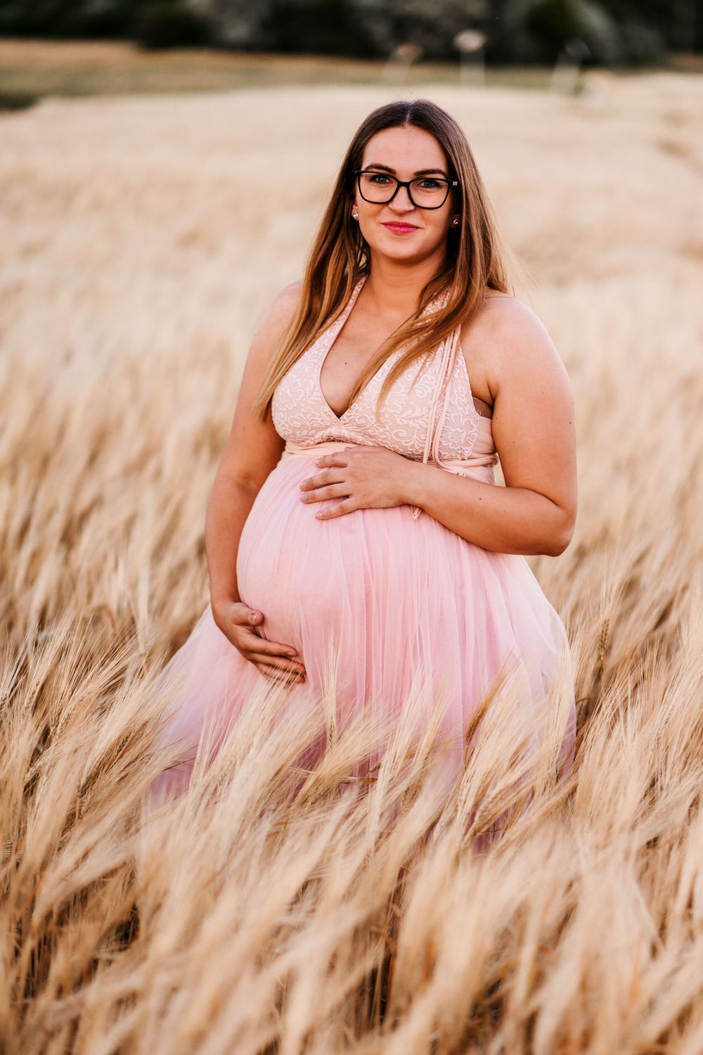 Expectant mother in a pink dress poses in a wheat field during golden hour.