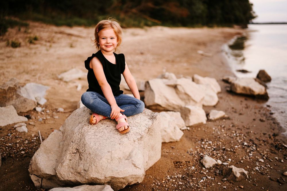 A child sits on a large rock by the water's edge during sunset.