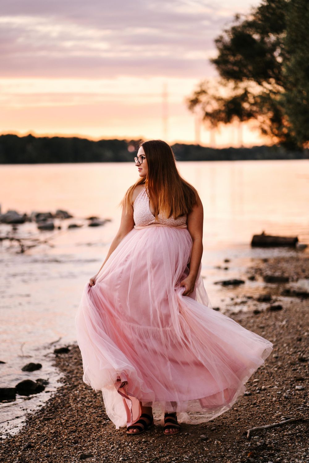Woman in pink tulle gown twirls on rocky shoreline at sunset.