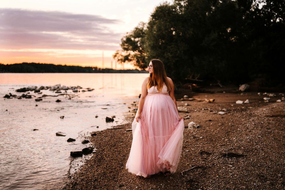 Woman in flowing pink dress walks along waterfront with rocks at sunset.