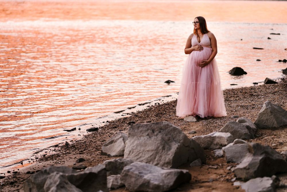 Maternity photo of woman in pink dress standing on rocky beach at sunset.