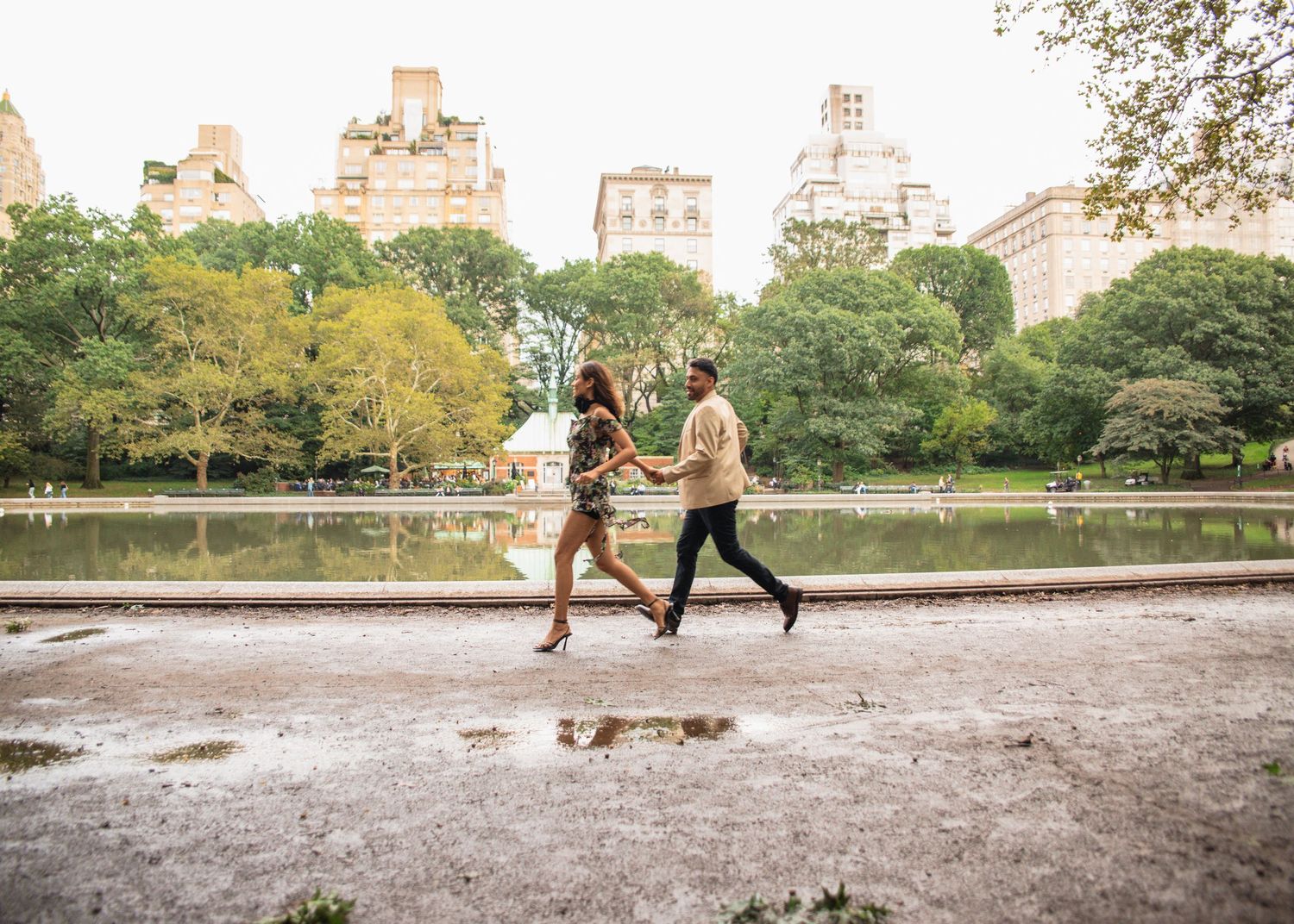 Young couple walks hand-in-hand along a rain-wet path beside a lake in Central Park, New York.