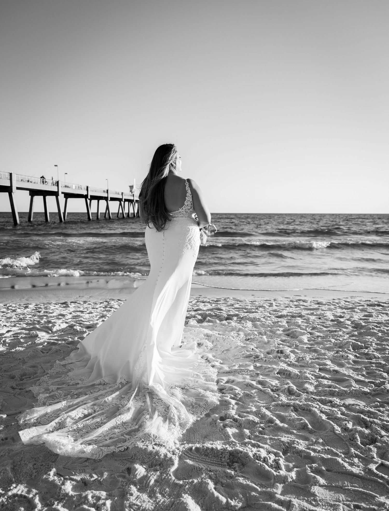 Black and white photo of bride in flowing dress walking along beach near pier.