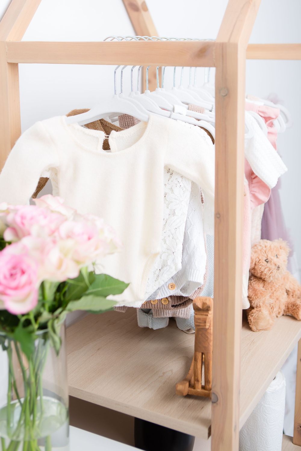 Delicate white baby clothes hanging on wooden rack with pink roses in vase.