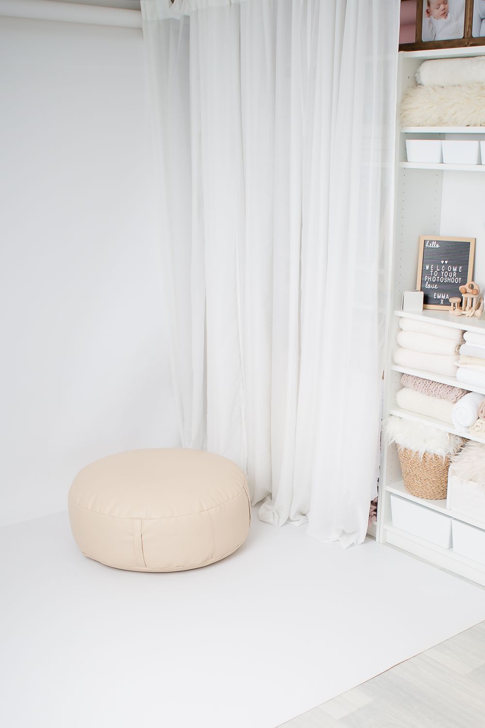 Beige round ottoman on white floor near white curtains and shelving unit.