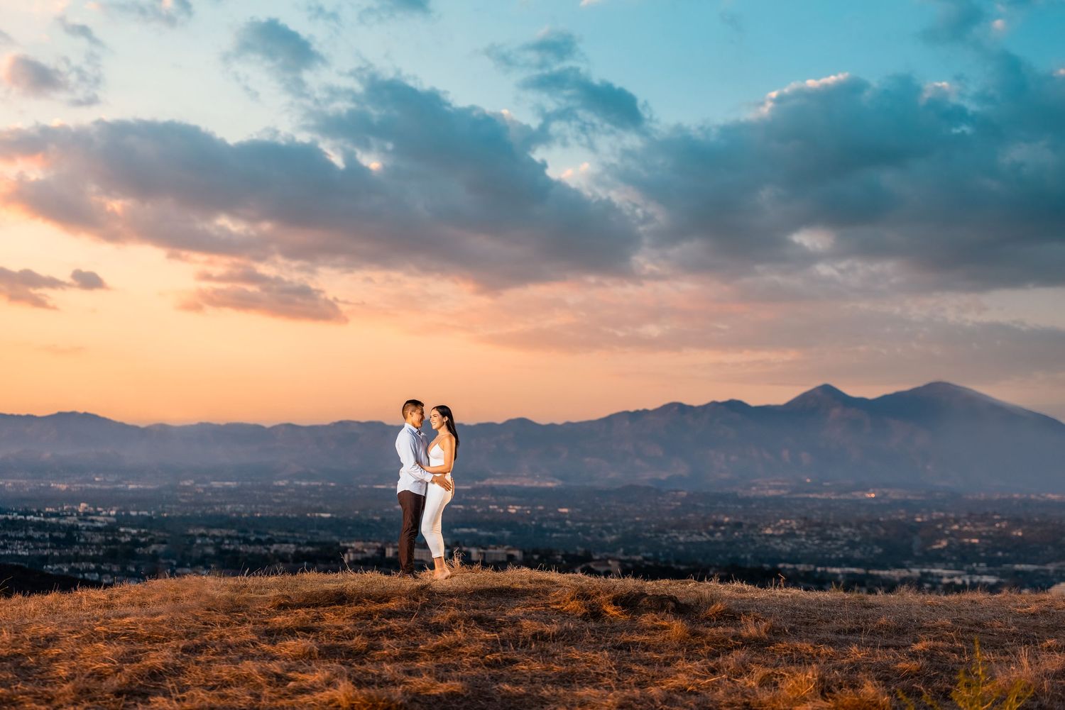 Scenic hilltop engagement portrait with mountain range and sunset sky in background.