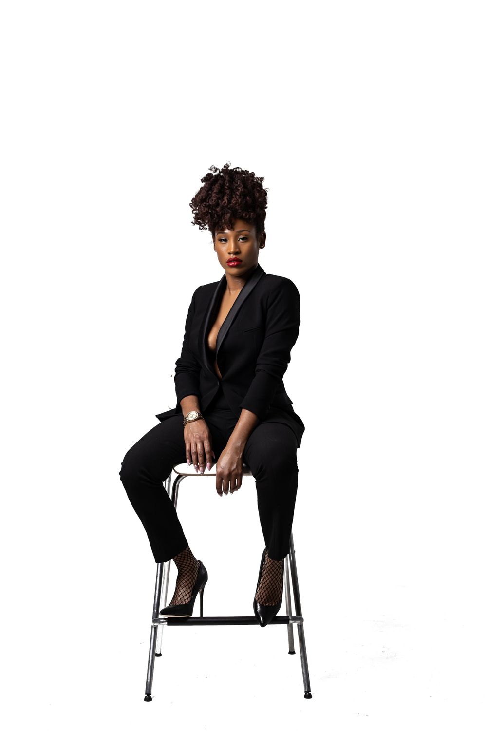 Elegant pose on a high stool wearing a tailored black suit against white background.