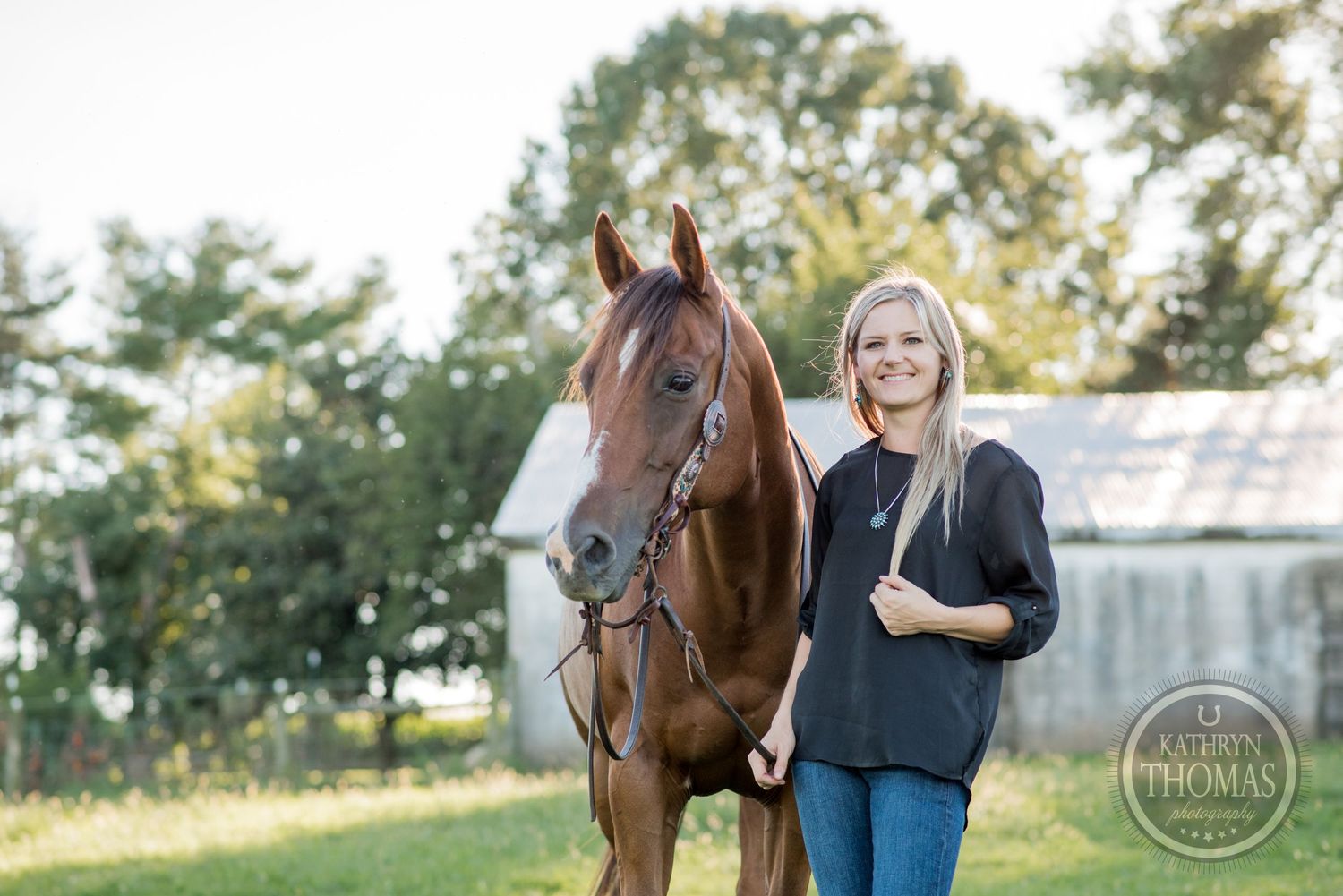 A chestnut horse with bridle poses with its handler in front of a white barn structure.