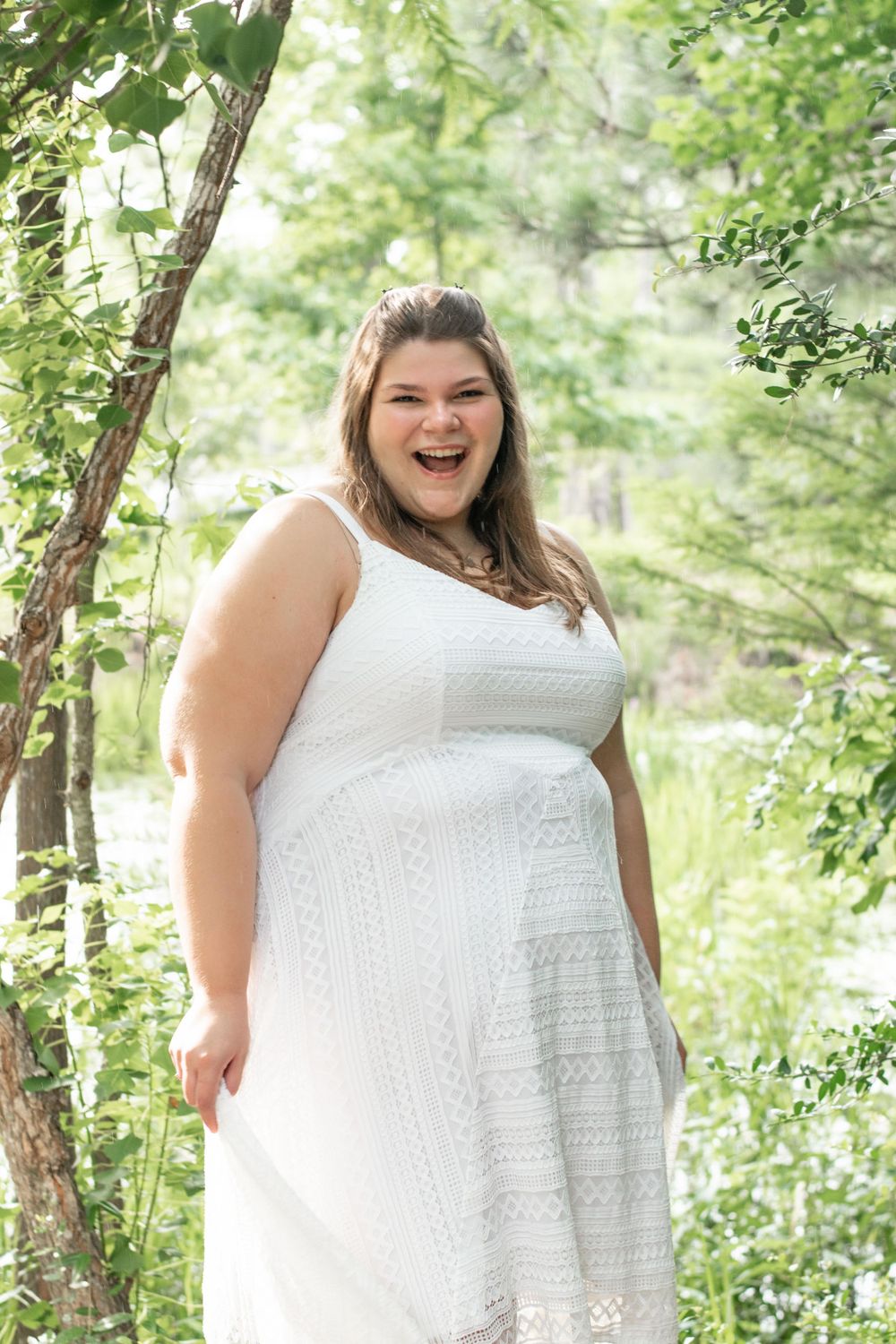 Portrait of someone in white lace dress standing among green forest foliage.