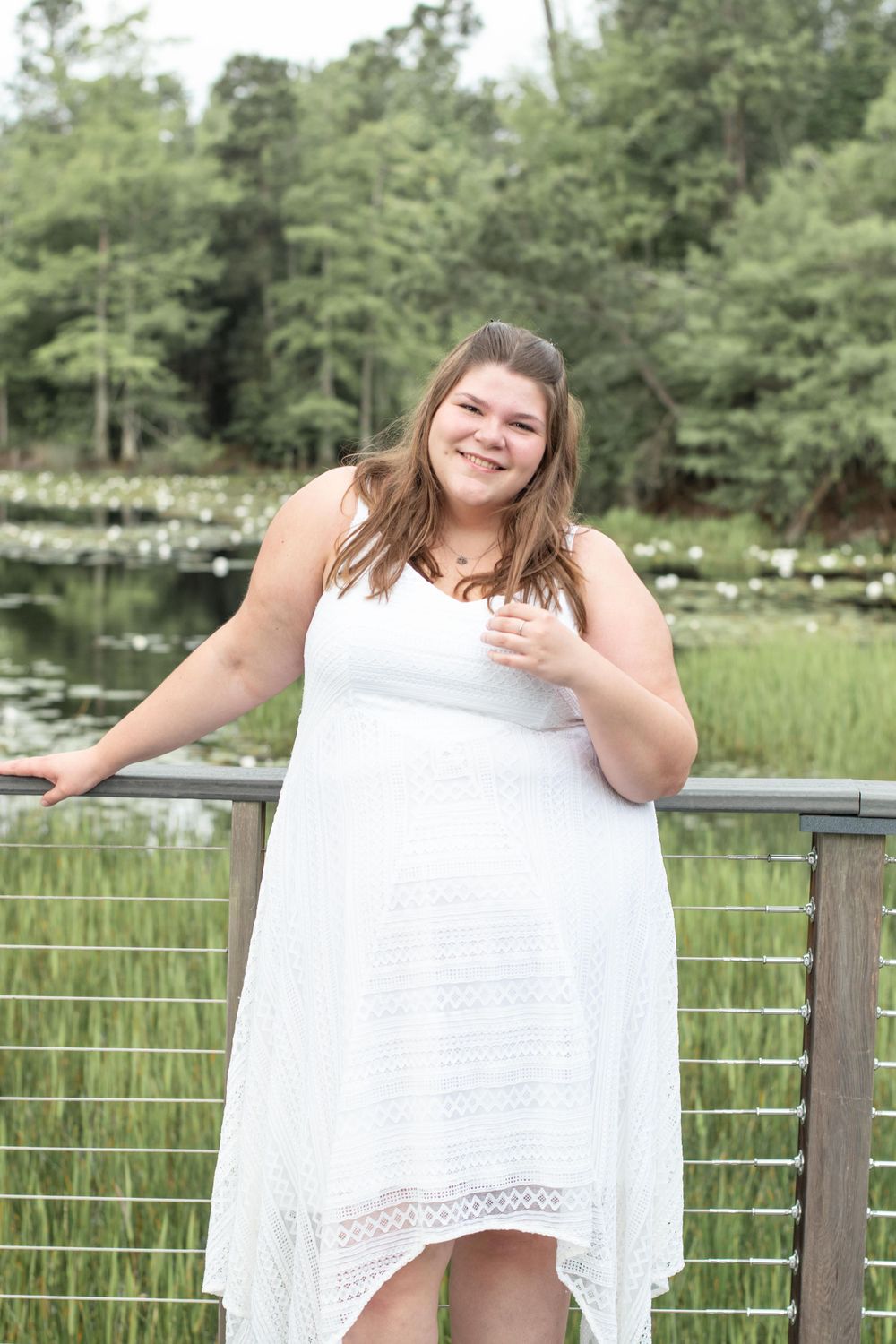 Someone in a flowing white dress poses joyfully on a boardwalk overlooking a pond with water lilies.