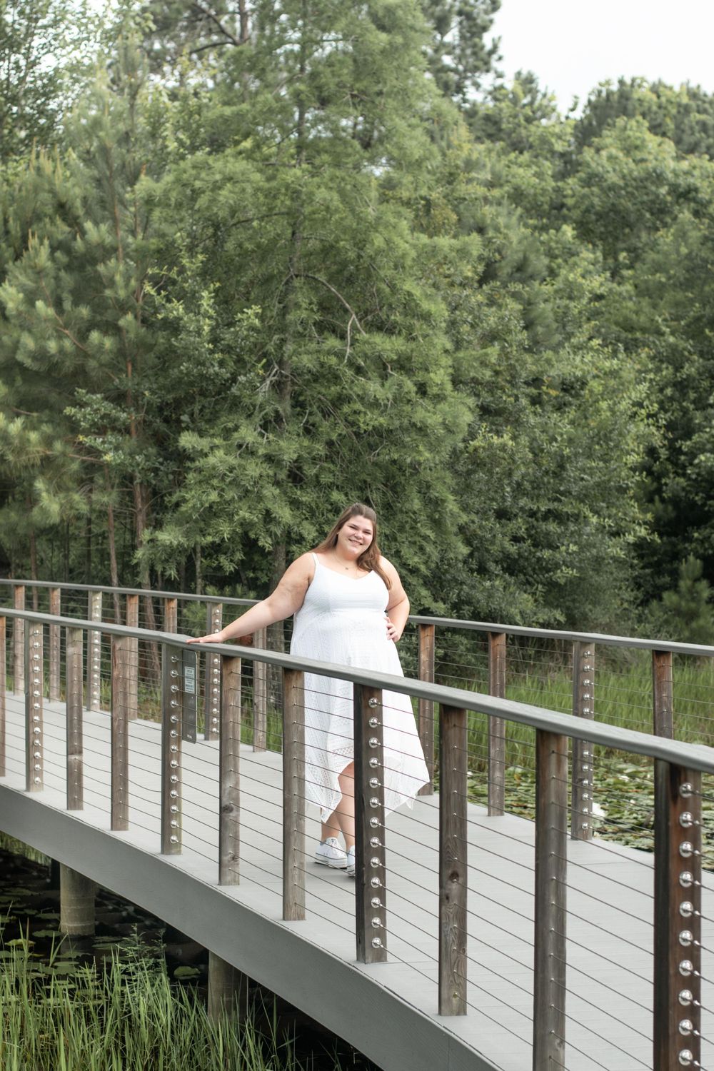Person in white dress strikes elegant pose on curved wooden bridge over pond.
