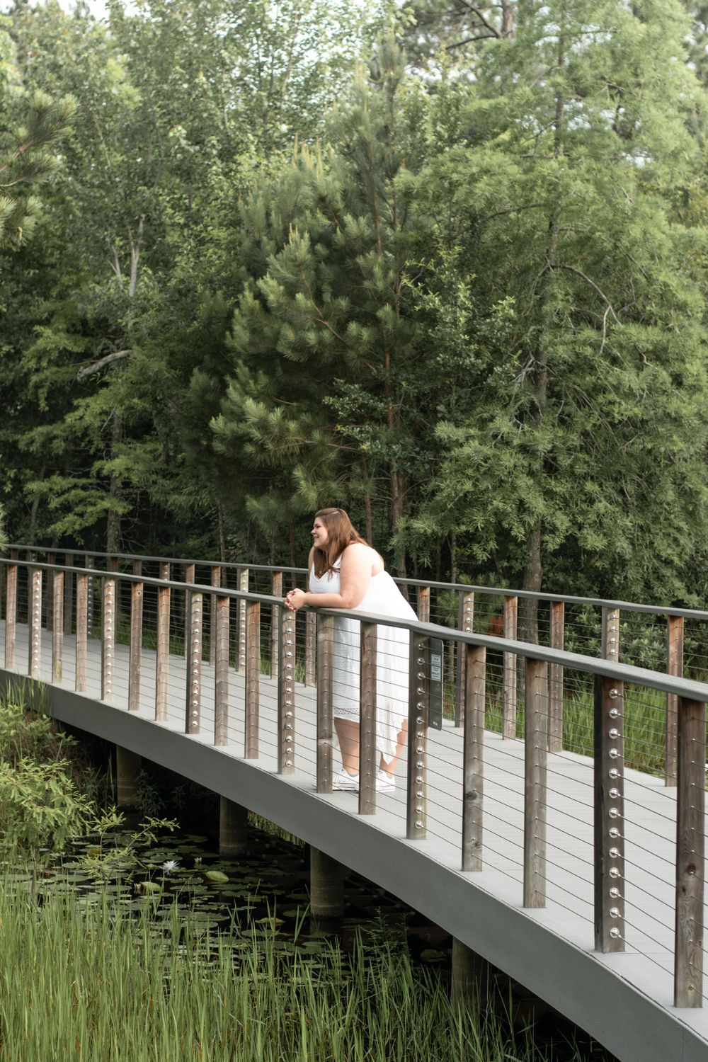 A person in white leaning on the railing of a curved wooden footbridge surrounded by pine trees and greenery.