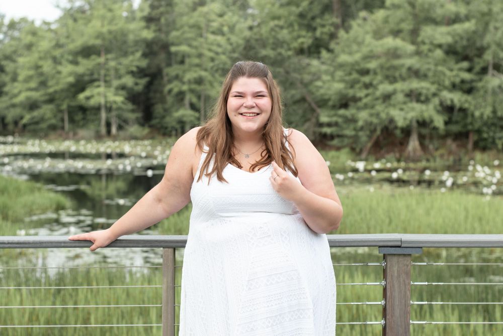 Someone in white dress poses happily on wooden boardwalk with pond and forest backdrop.