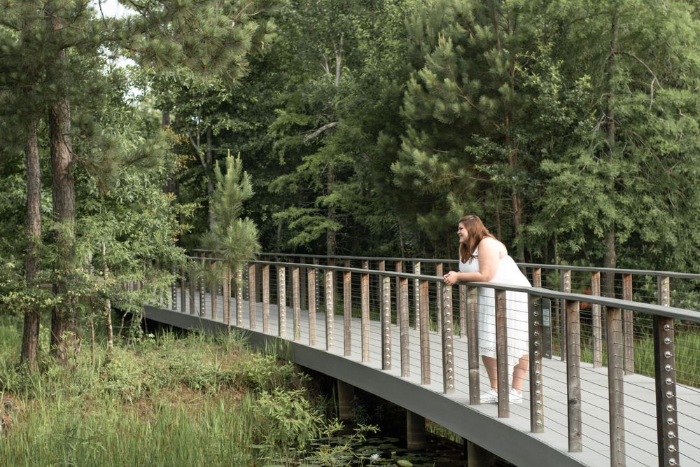 Person in white dress standing thoughtfully on wooden bridge surrounded by lush forest scenery.