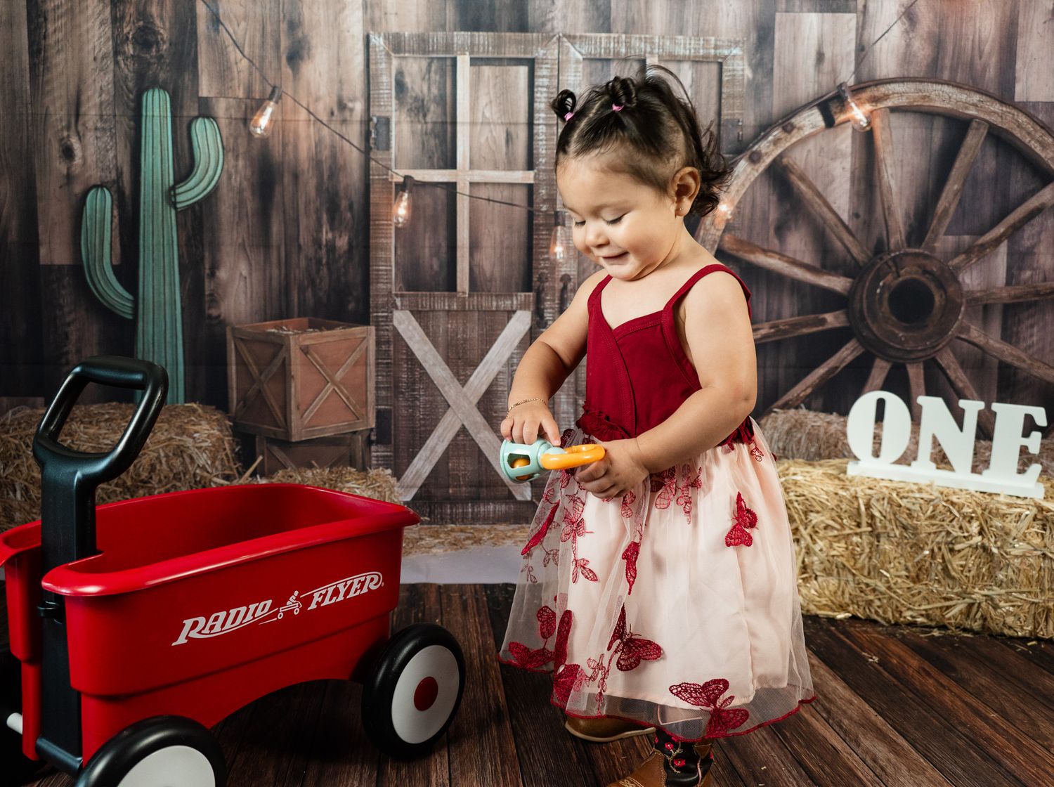 Child in red and white dress plays near vintage wagon and rustic western-themed backdrop.