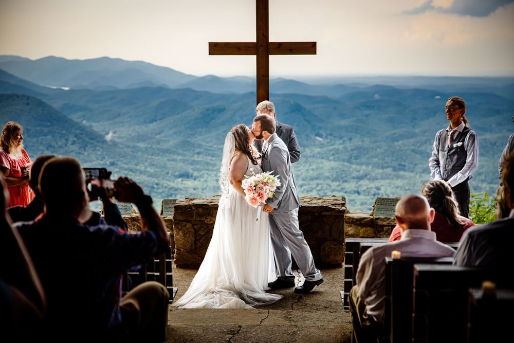 A scenic mountaintop wedding ceremony taking place in front of a large wooden cross with guests seated outdoors.