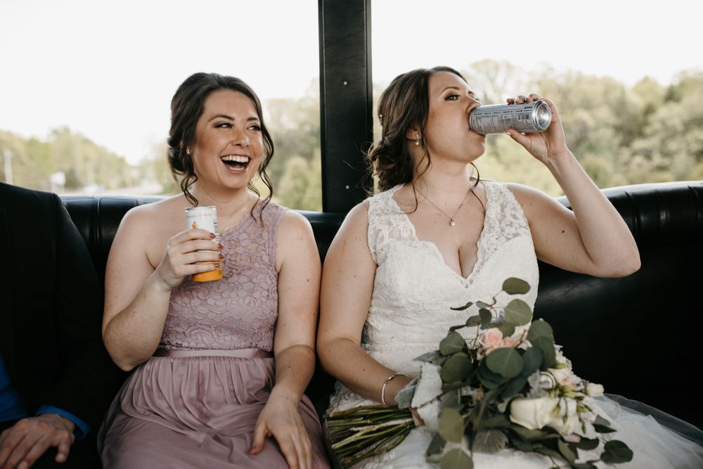Bride drinking from can while bridesmaid laughs on party bus.