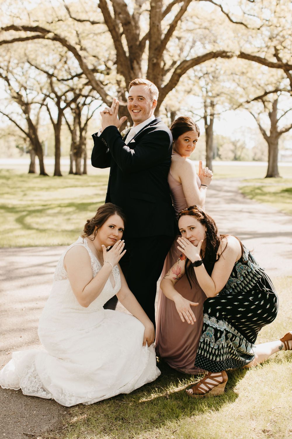Bride, groom, and guests posing playfully outdoors under large trees.