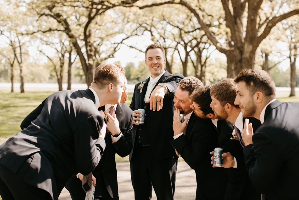 Groom shows off wedding ring to laughing groomsmen in black suits outdoors among spring trees.