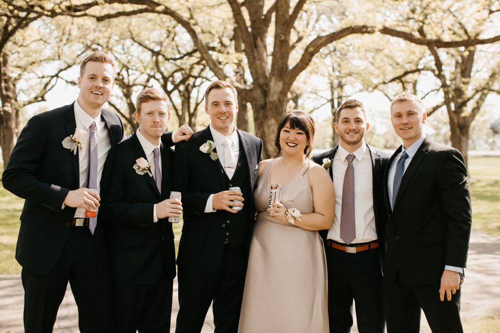 Smiling wedding party group poses outdoors in black suits and blush dress holding drinks by trees.