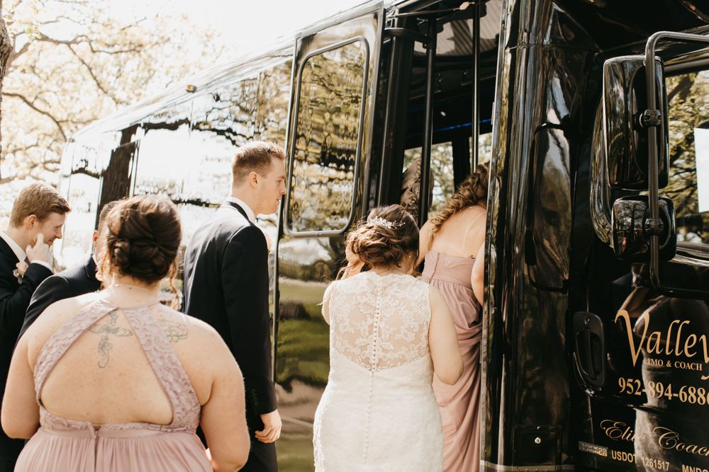 Wedding party boards black party bus with bride in white gown and bridesmaids in blush dresses.