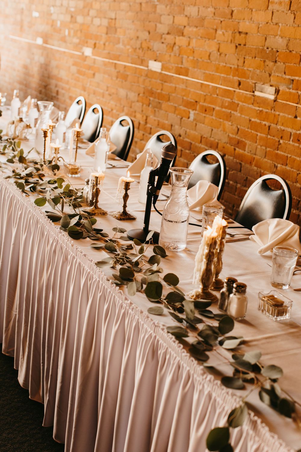 Elegant wedding head table with eucalyptus garland, candles, and skirted linens against brick wall.