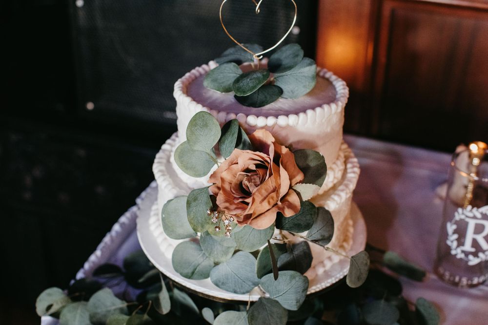 Two-tier white wedding cake with rose, eucalyptus leaves, and gold heart topper closeup.