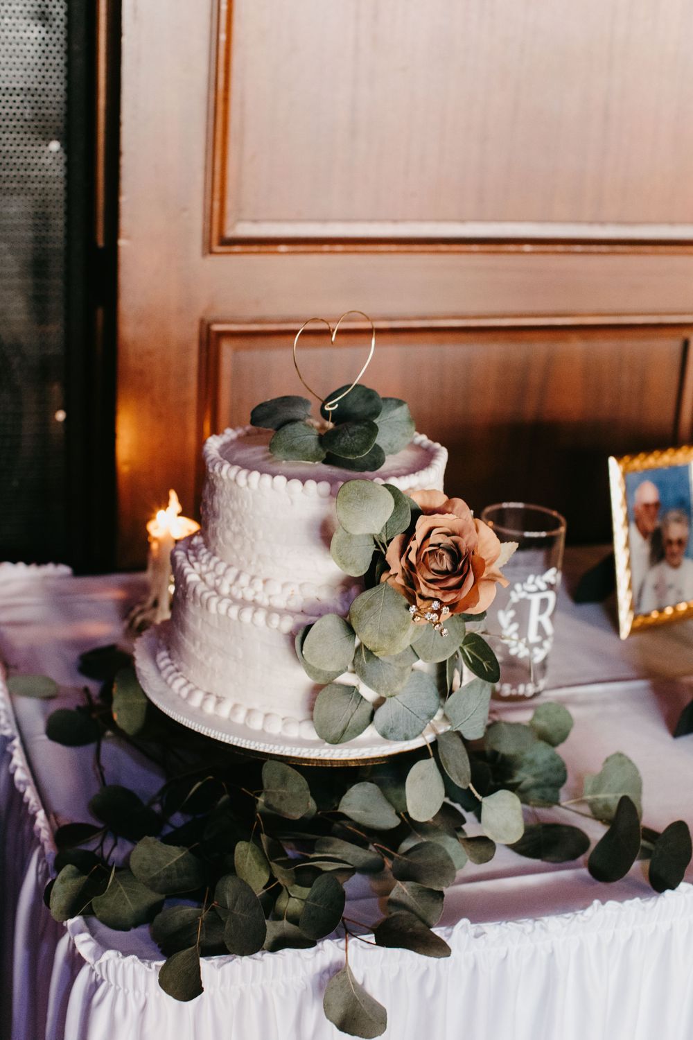 Two-tier white wedding cake decorated with rose, eucalyptus, and gold heart topper on draped table.