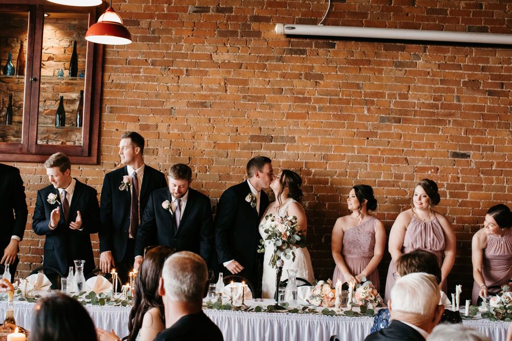 Bride and groom share a kiss at head table during wedding reception in brick-walled venue.