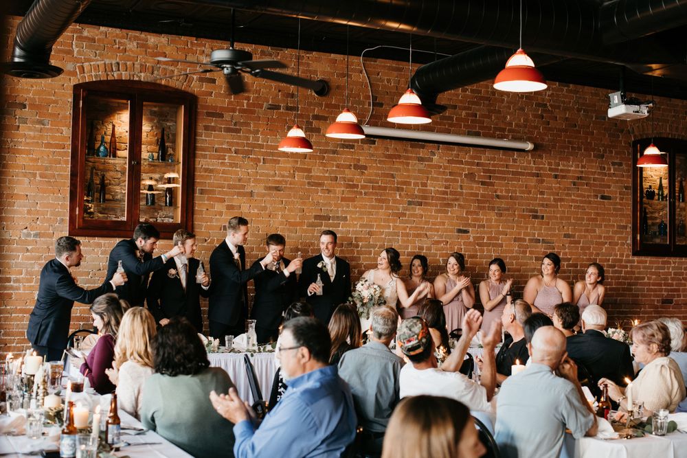 Wedding party stands at head table during reception toasts in rustic brick-walled venue with guests.