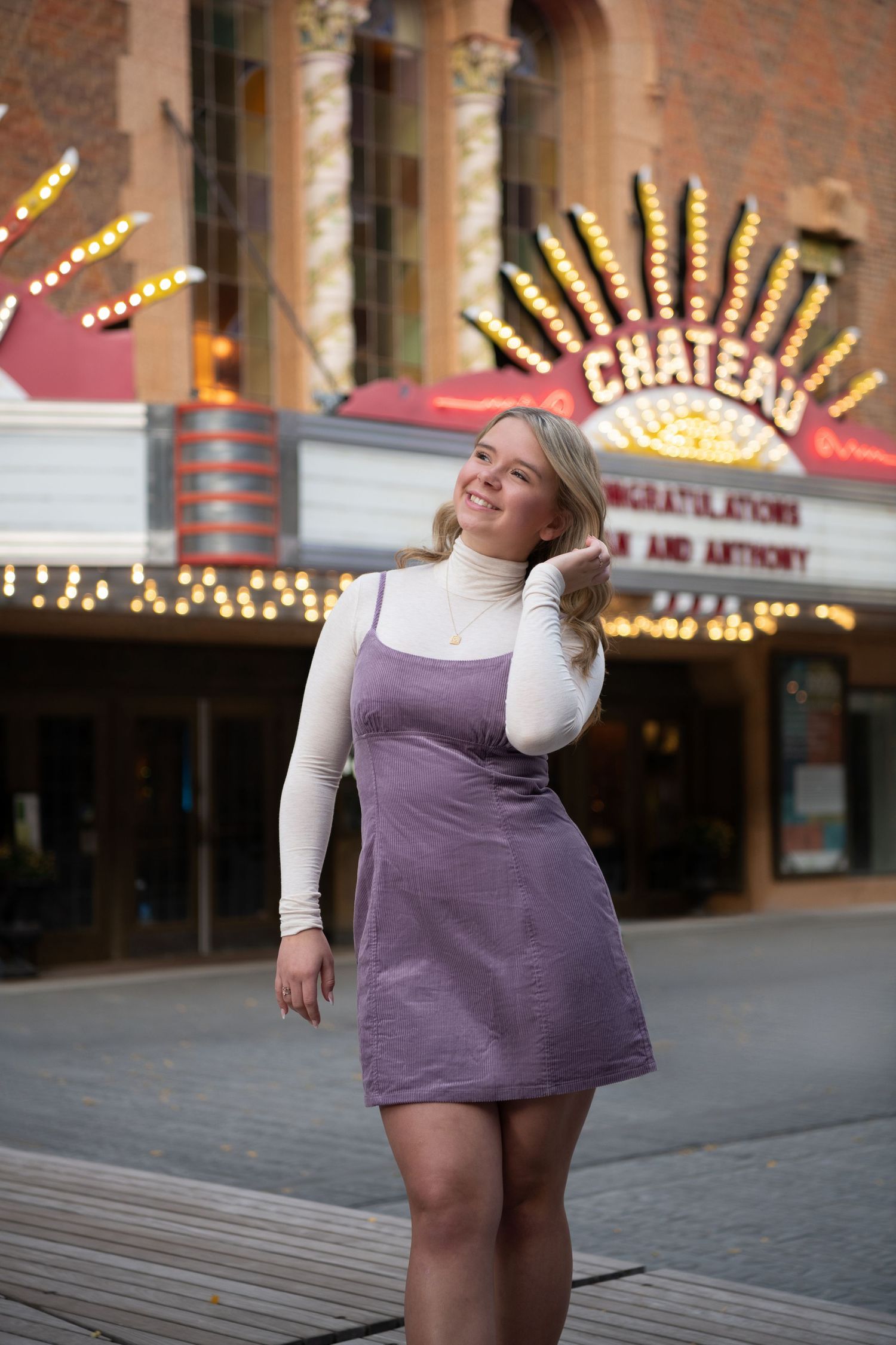 Fashion portrait of figure in purple dress and white turtleneck standing outside theater with illuminated marquee.