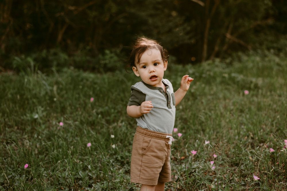 Young toddler in gray shirt and brown shorts stands expressively in an outdoor setting.