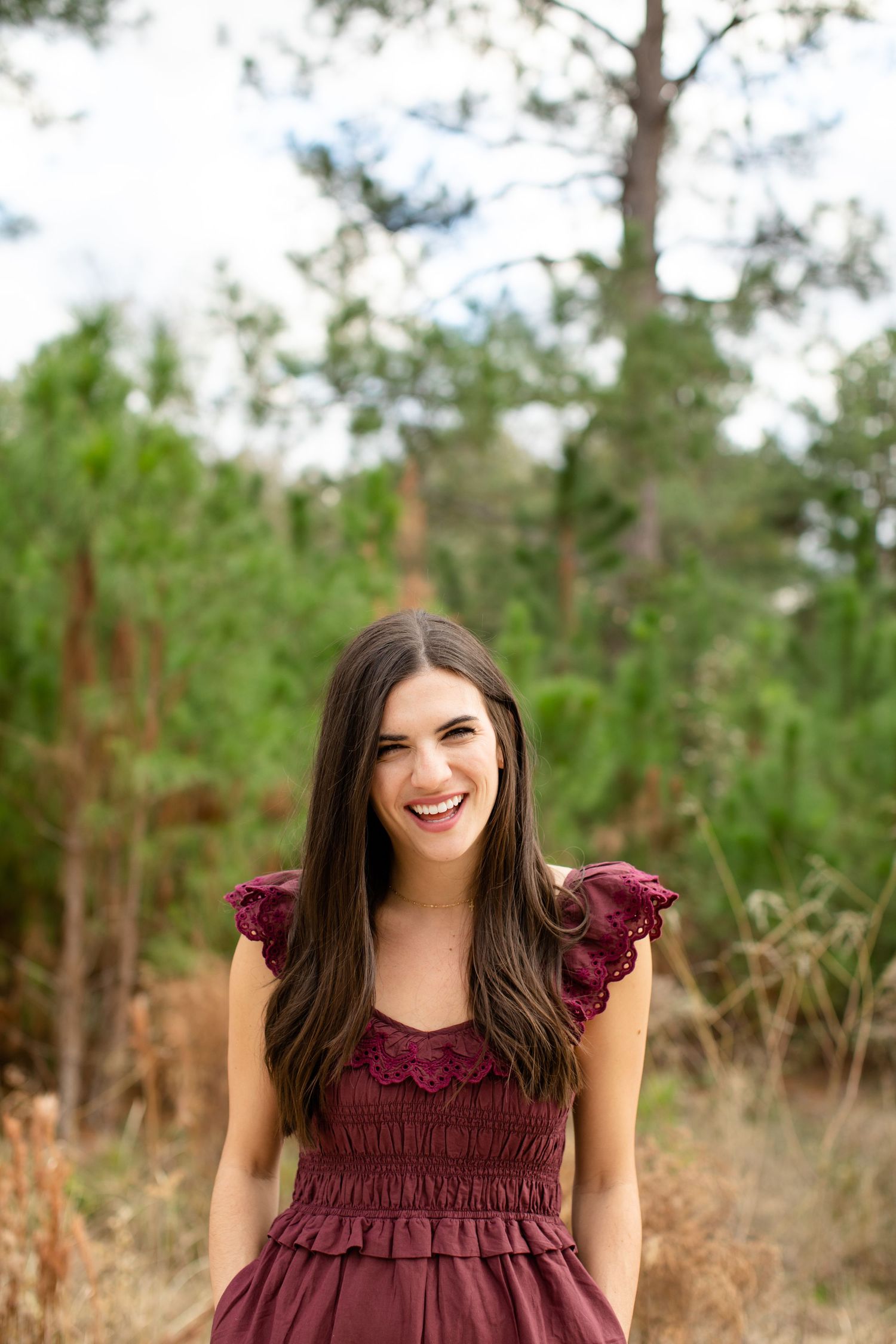 Woman in burgundy ruffle dress smiles brightly against pine forest background.