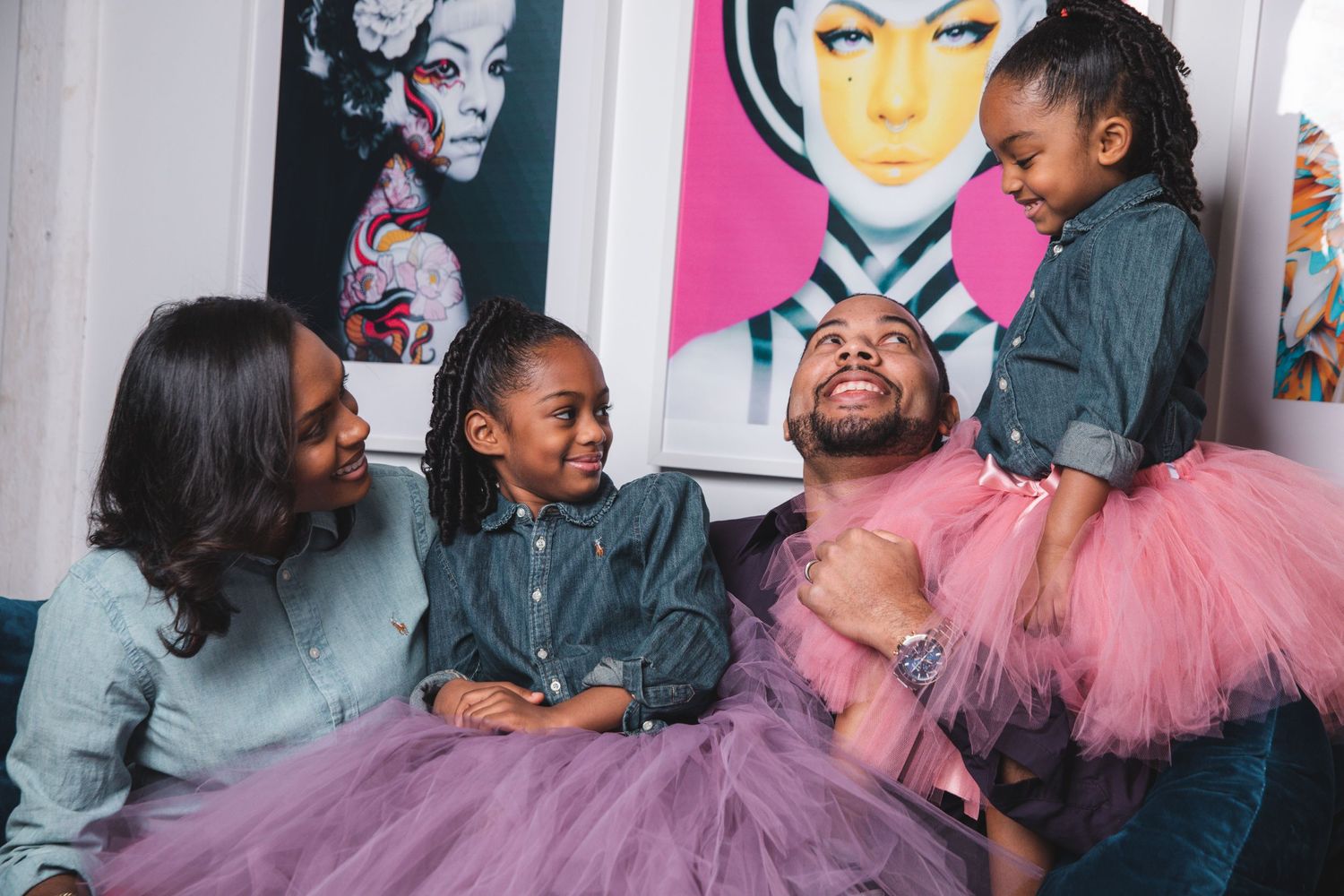 Joyful Black family laughing together on couch wearing matching denim and pink tutu outfits in colorful room.