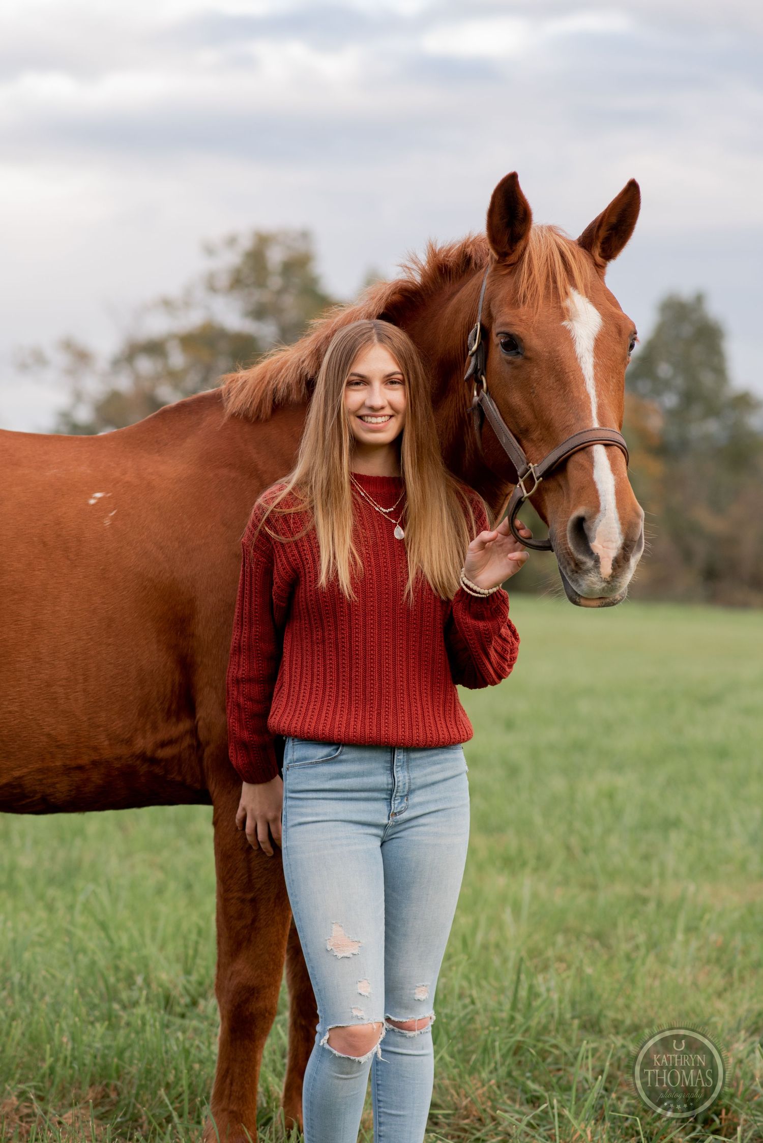 A chestnut horse with bridle stands close to someone in a red knit sweater and distressed jeans.