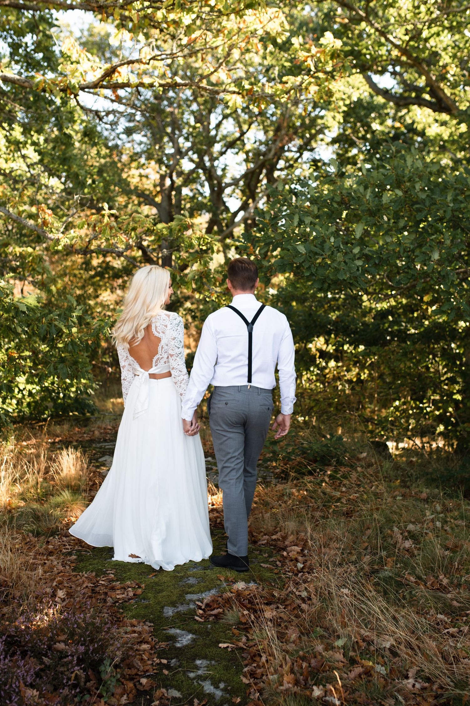 A wedding couple walks hand in hand through a sun-dappled forest path in elegant attire.