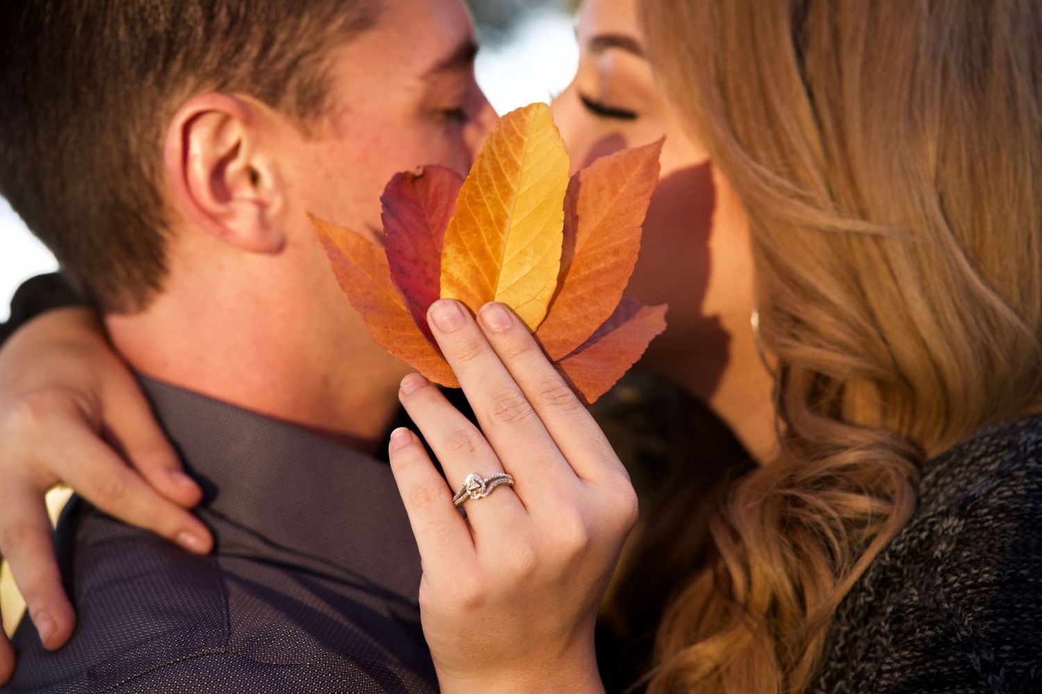 Romantic autumn scene with a couple holding leaves to hide a kiss and showing sparkling engagement ring.
