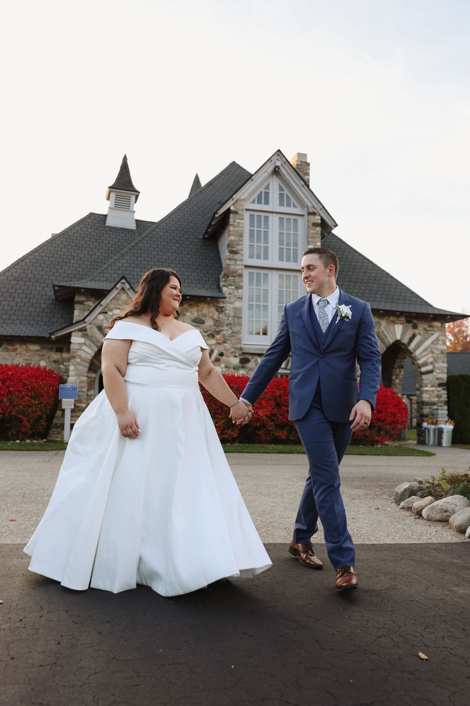 A newlywed couple in wedding attire holding hands and walking together outside a stone church with red flowers.