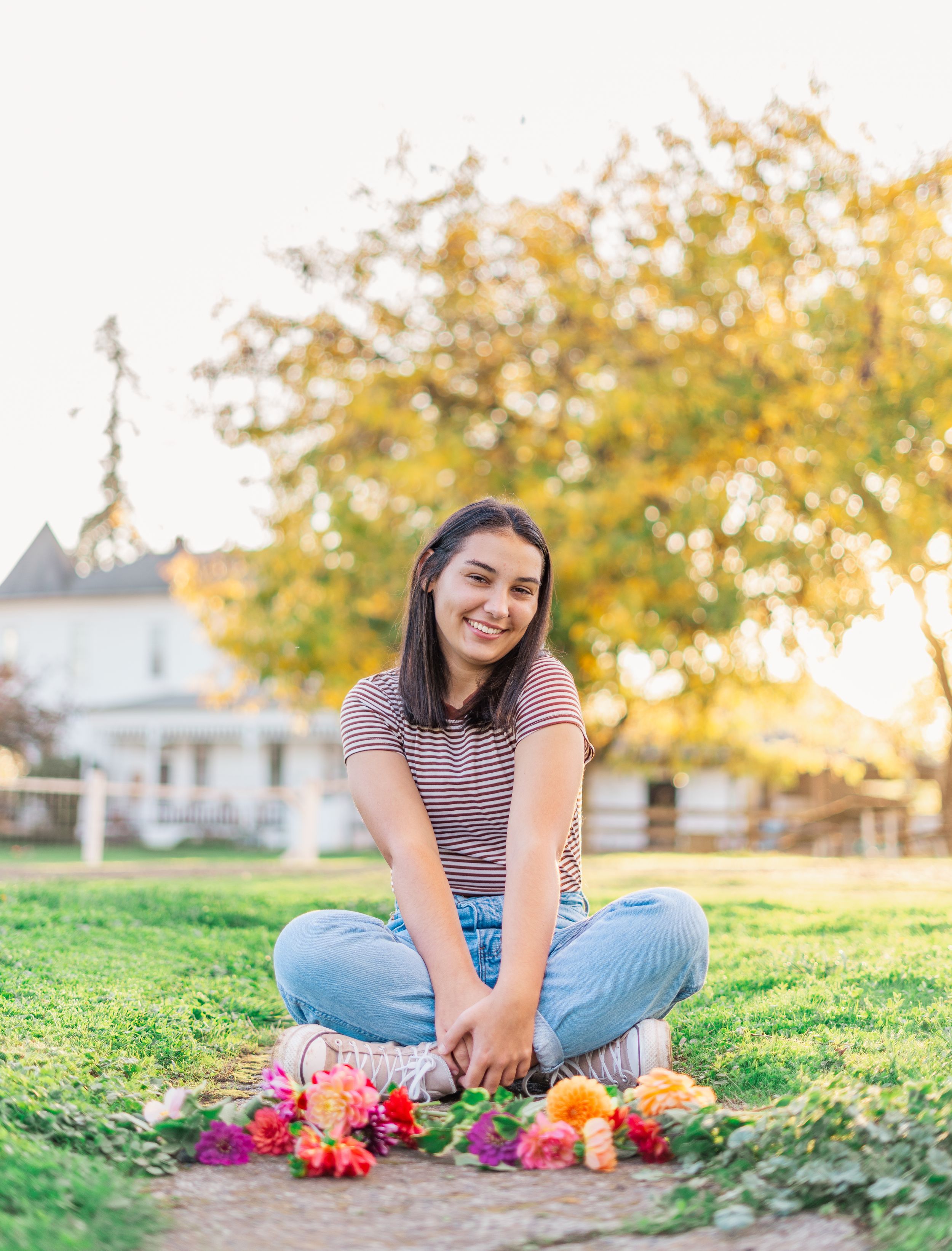 A high school senior sits cross-legged on grass with colorful flowers while smiling on a sunny autumn day.