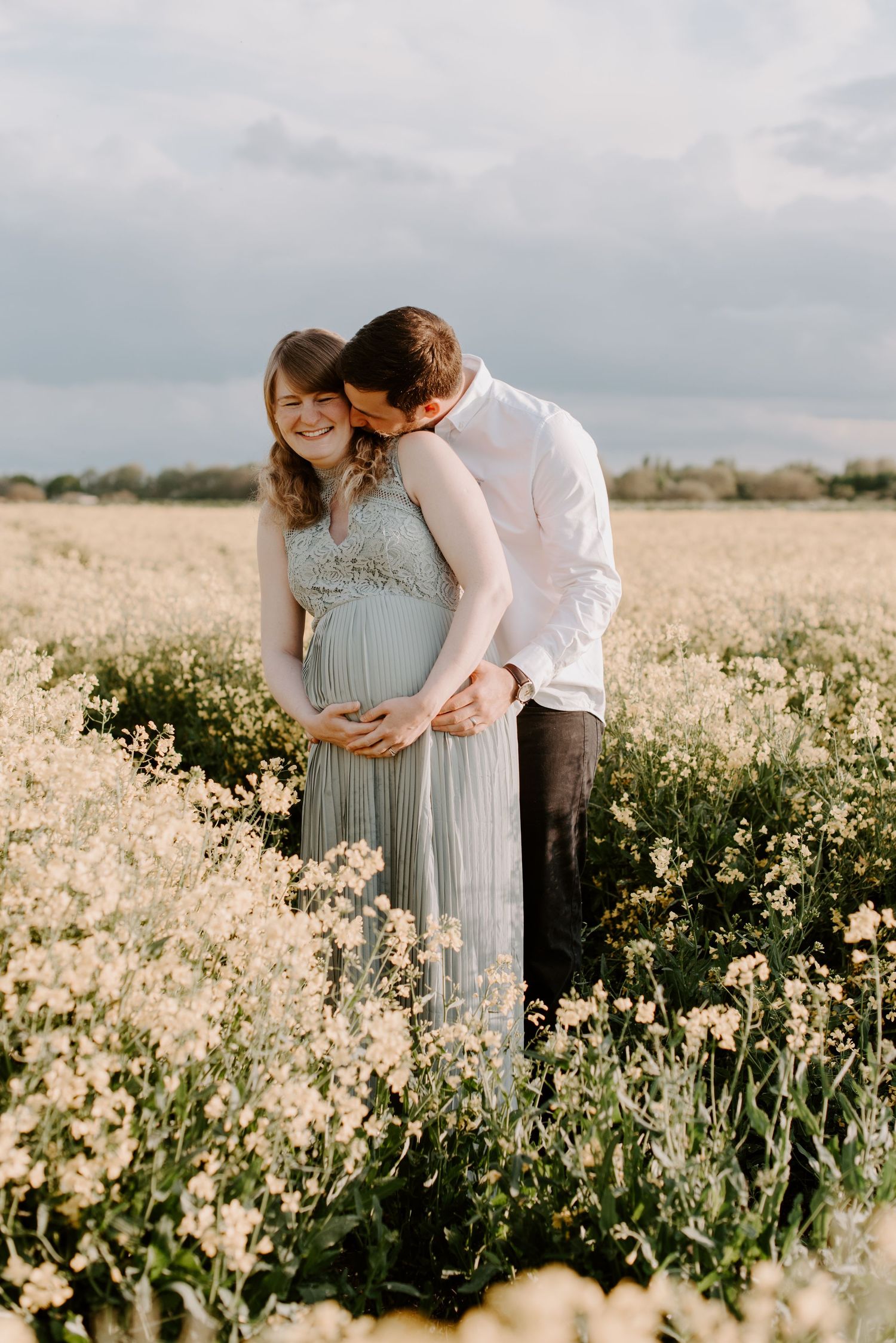 A pregnant lady is holding her bump. Her husband stands behind her and is kissing her neck, making her laugh.